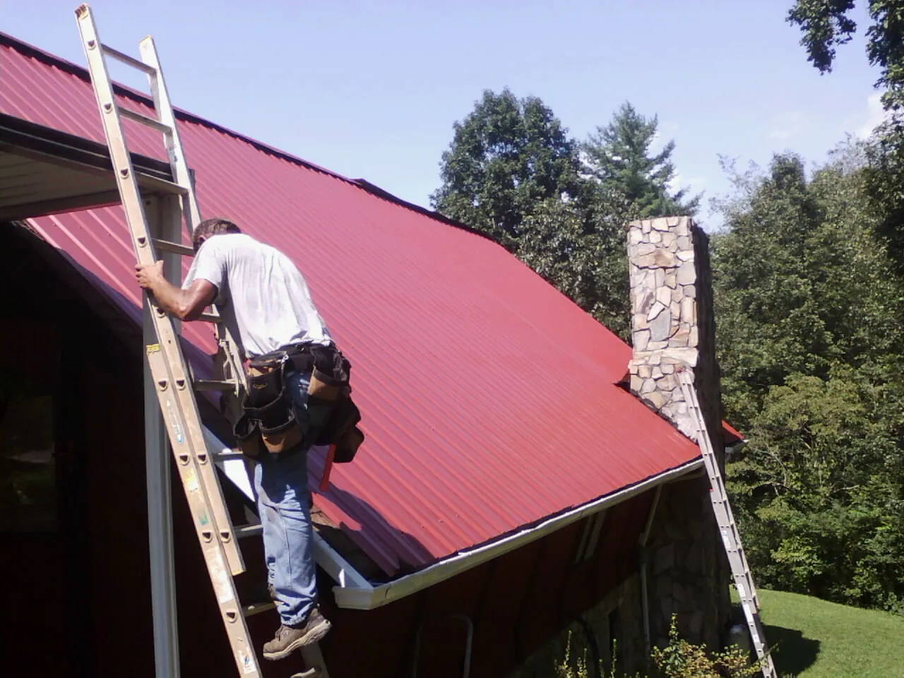 Roofer on a ladder beside a red metal roof and stone chimney — Soffit & Fascia Repair by Roofing Brothers in Homerville